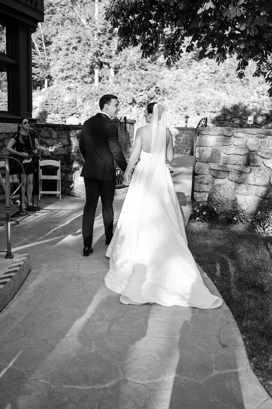 Black and white film photo of the couple walking hand-in-hand down a stone path while looking at each other.